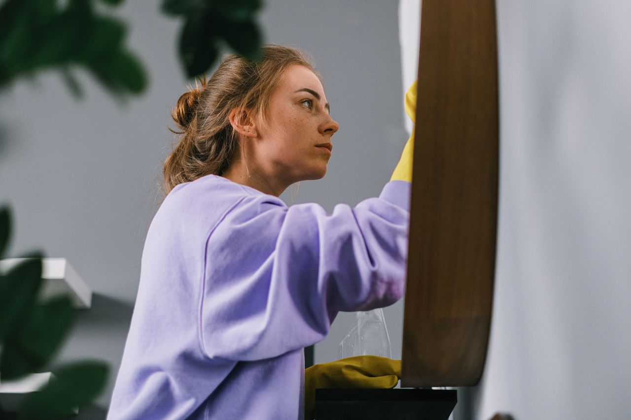 Side view of young focused female in latex gloves standing and cleaning mirror in light room in daytime